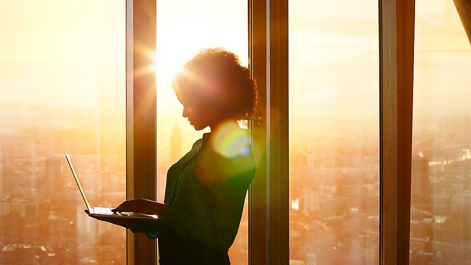 Une femme debout utilise un ordinateur portable sur fond de coucher de soleil derrière les fenêtres d’un immeuble