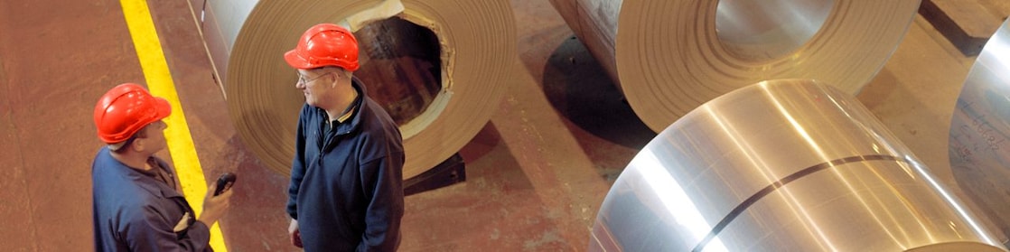 Two factory workers standing next to rolls of steel