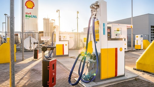 Une station de ravitaillement Shell BioLNG avec plusieurs distributeurs de GNL aux couleurs jaune et blanc de Shell. Des tuyaux sont connectés aux pompes, et des réservoirs de stockage ainsi que des équipements industriels apparaissent à l’arrière‑plan sous une lumière intense.