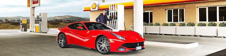 Une Ferrari rouge arrêtée à la pompe d'une station Shell avec un homme qui s'apprête à faire le plein