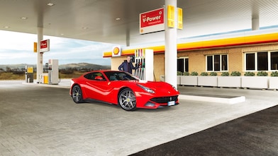 Une Ferrari rouge arrêtée à la pompe d'une station Shell avec un homme qui s'apprête à faire le plein