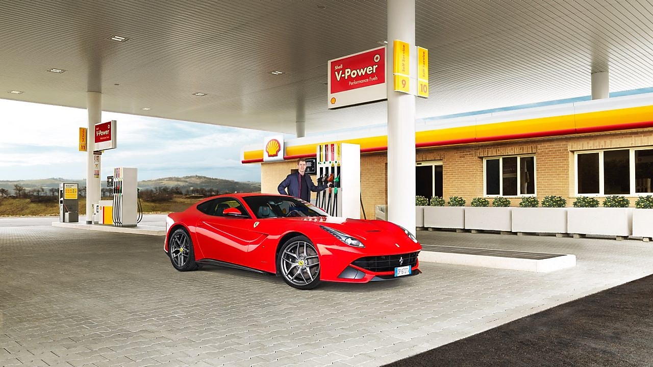 Une Ferrari rouge arrêtée à la pompe d'une station Shell avec un homme qui s'apprête à faire le plein
