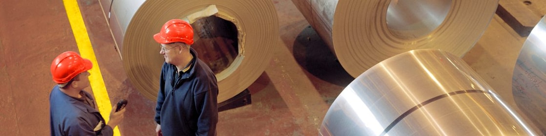 Two factory workers standing next to rolls of steel