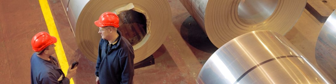 Two factory workers standing next to rolls of steel