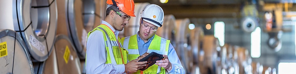 2 mannen in veiligheidsjassen en helmen kijken naar een tablet, op elkaar gestapeld industrieel materiaal op de achtergrond