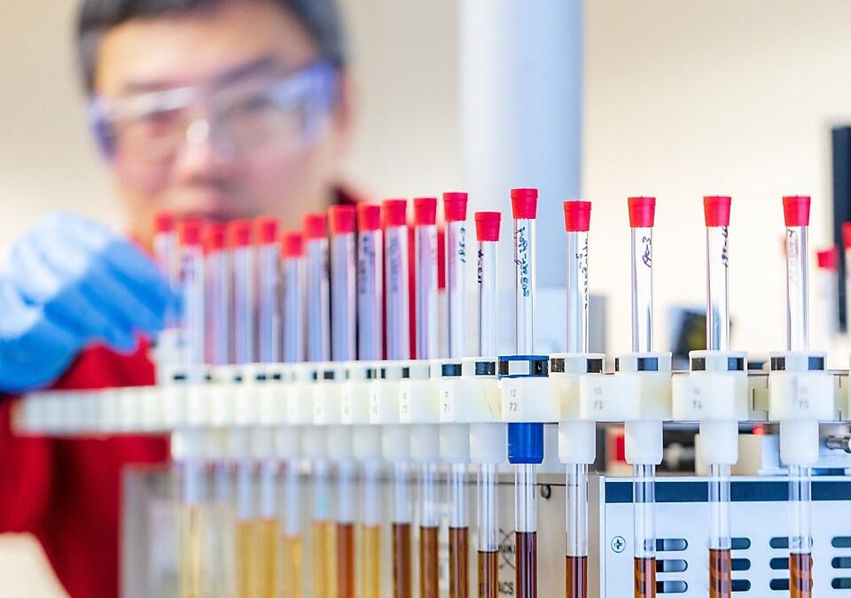 Man in rode laboratoriumjas en blauwe handschoenen, bezig met reageerbuizen met vloeistofmonsters in een laboratoriumomgeving.