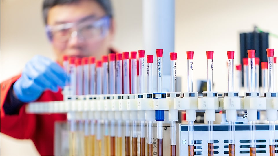 Man in rode laboratoriumjas en blauwe handschoenen, bezig met reageerbuizen met vloeistofmonsters in een laboratoriumomgeving.