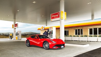 Een rode Ferrari staat aan een Shell-takstation geparkeerd met een man die leunt op een benzinepomp