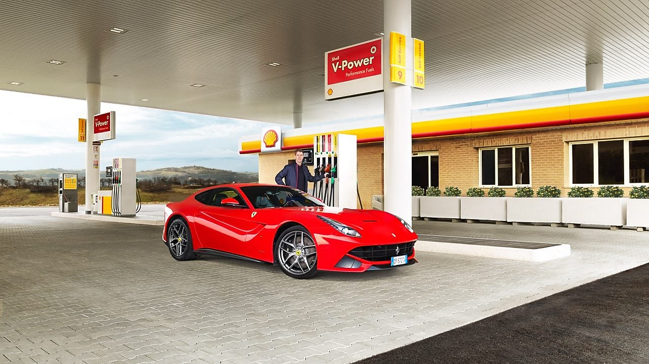 Een rode Ferrari staat aan een Shell-takstation geparkeerd met een man die leunt op een benzinepomp