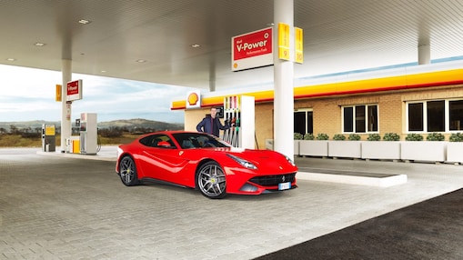 Een rode Ferrari staat aan een Shell-takstation geparkeerd met een man die leunt op een benzinepomp