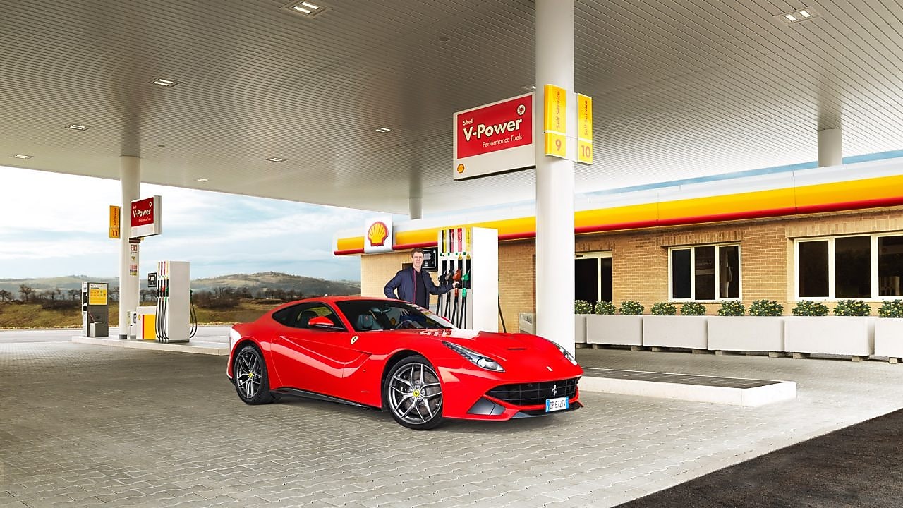 Een rode Ferrari staat aan een Shell-takstation geparkeerd met een man die leunt op een benzinepomp