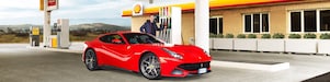 Een rode Ferrari staat aan een Shell-takstation geparkeerd met een man die leunt op een benzinepomp