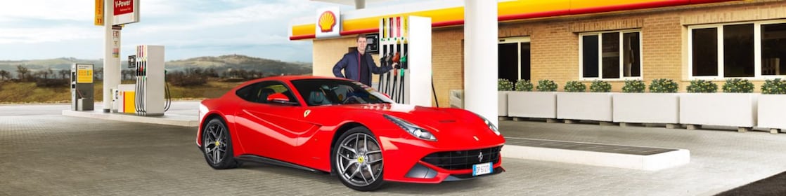 Een rode Ferrari staat aan een Shell-takstation geparkeerd met een man die leunt op een benzinepomp