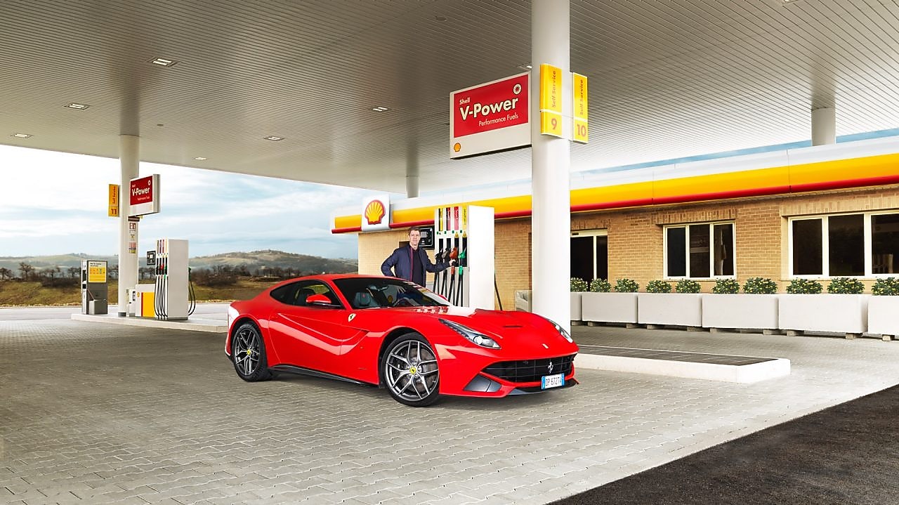 Een rode Ferrari staat aan een Shell-takstation geparkeerd met een man die leunt op een benzinepomp