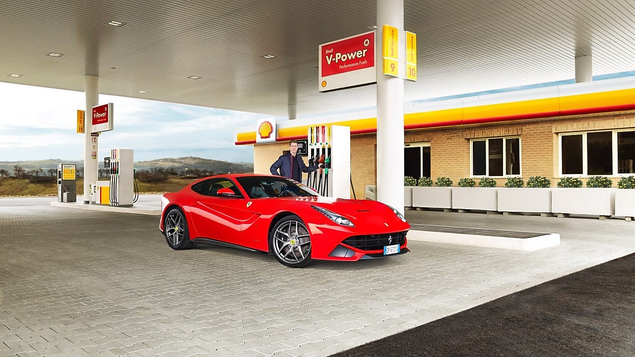 Een rode Ferrari staat aan een Shell-takstation geparkeerd met een man die leunt op een benzinepomp