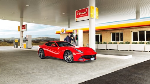Een rode Ferrari staat aan een Shell-takstation geparkeerd met een man die leunt op een benzinepomp